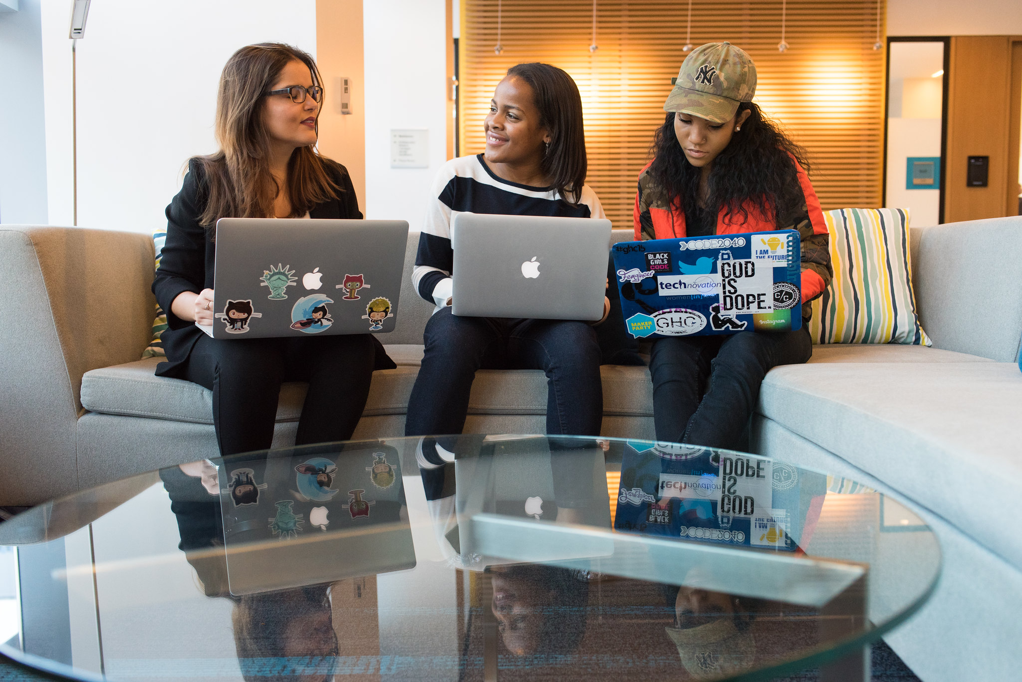 Three women of color sitting on a couch, each with a laptop covered, some of them adorned with coding-related stickers